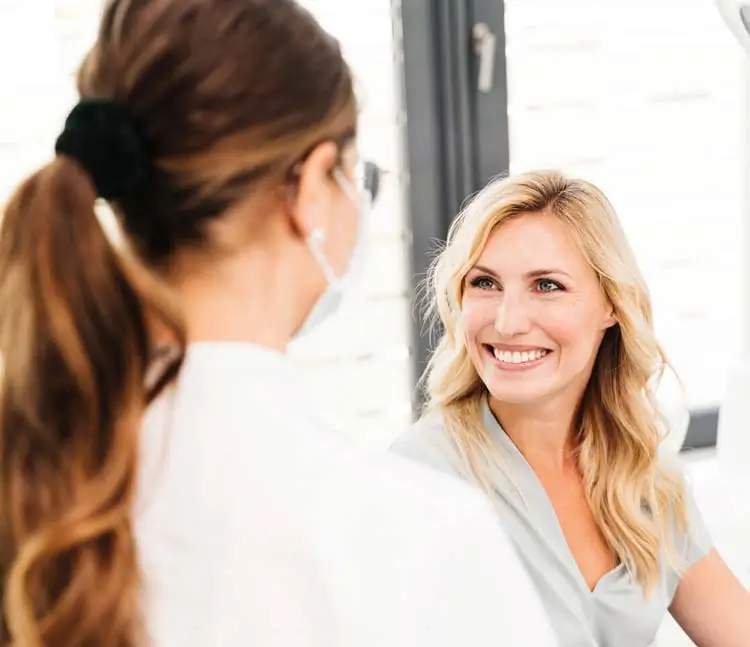 Blick &uuml;ber die Schulter einer Zahn&auml;rztin auf eine l&auml;chelnde Patientin 