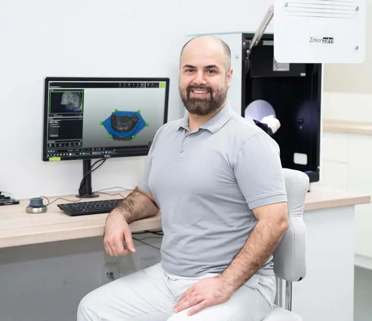 Labortechniker sitzt an einem Scanner mit dem dazugehörigen 3D-Modell am PC und lächelt in die Kamera