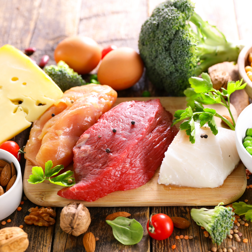 Rohe Fleischst&uuml;cke, H&auml;hnchenbrust, Fischfilet, Eier, Brokkoli, K&auml;se, N&uuml;sse und Kirschtomaten auf Holzbrett und Tisch verteilt
