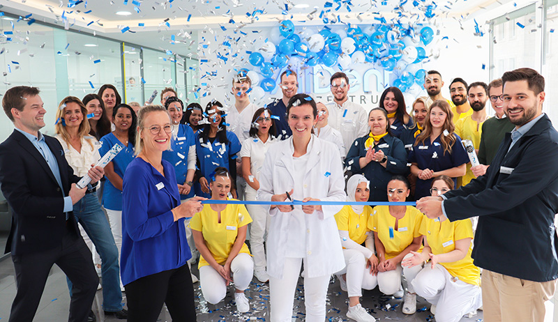 Menschen in bunter Arbeitskleidung schneiden ein blaues Band durch, w&auml;hrend blaue Luftballons und Konfetti fallen.